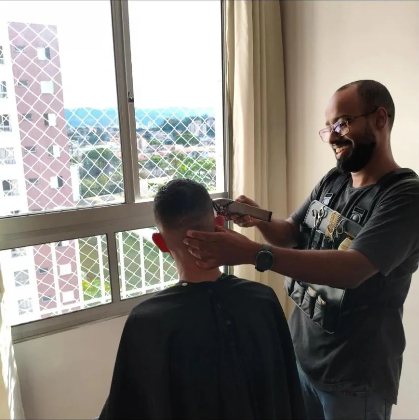 Eliabe Mayer, sorrindo, cortando o cabelo de um cliente de costas, perto da janela de um apartamento